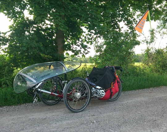 ein Liegedreirad mit durchsichtigem Wind- und Regenschutz steht auf einem ungeteerten Weg. Lange orange Fahne dran