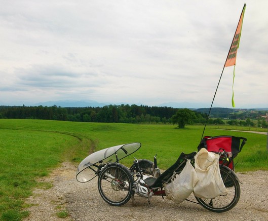 ein Liegedreirad mit durchsichtigem Wind- und Regenschutz steht auf einem ungeteerten Weg. Lange orange Fahne dran. Blick auf eine weite Wiese. seitlich am Rad hängen beige ehemalige Sandsäcke
