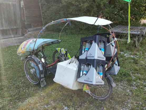 Das Liegedreirad mit Wind- und Regenschutz und Dach beladen mit vier leeren Wasserkästen und noch seitlich einer Tasche mit Leergut