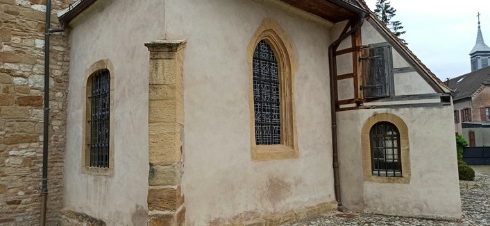 Bild zeigt einen Seitenanbau an einer Kirche mit zwei Fenstern, das linke mit Rundbogen oben, das rechte mit Spitze gotisch. das rechte ist aus buntem Bleiglas, man sieht die Stege zwischen den Glaselementen. Der Anbau ist weiß verputzt, an der Ecke eine Säule aus hellem Sandstein. An den Anbeu schließt ein Stück Fachwerkgebäude mit zwei weiteren Fenstern an, oben aus Holz, unten vergittert mit Rundbogen.