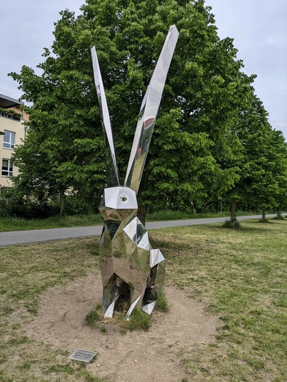 ein riesiger verchromter Hase mit extra langen Ohren, dessen Oberfläche aus lauter glatten reflektierenden Flächen zusammen gesetzt ist, sitzt im Rasen. Dahinter Laubbäume 