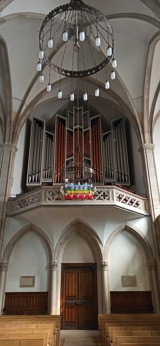 Orgel auf gotischer Empore. Prospekt eingefaßt mit weißem Gehäuse, Pfeifen in den inneren vier Feldern aus Kupfer.
Am Emporengeländer hängt die Regenbogenflagge mit dem Schriftzug PACE.