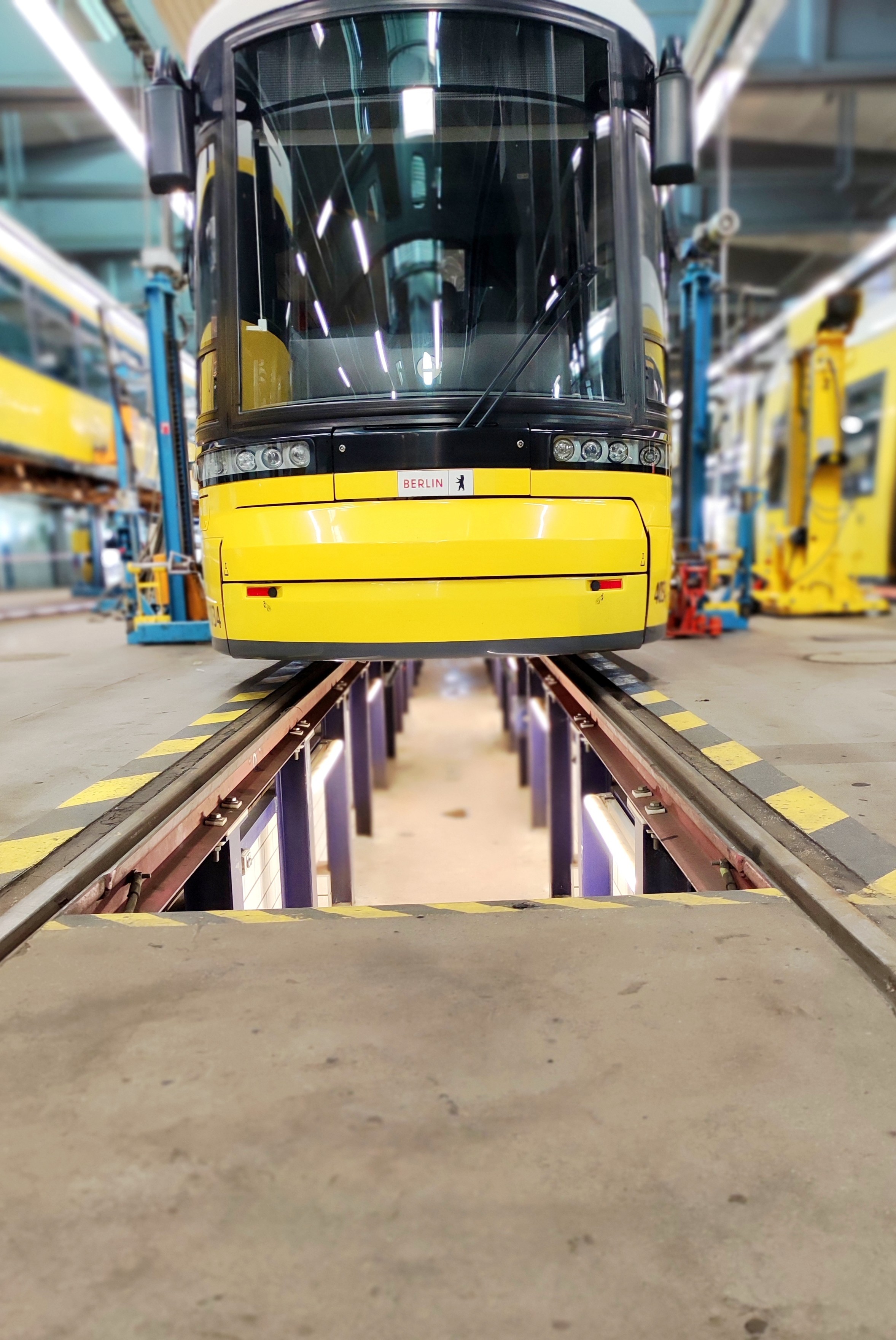Das Bild zeigt eine gelbe Straßenbahn, die auf einem Gleis in einer Werkshalle steht. Die Straßenbahn ist mit dem Schriftzug "BERLIN" gekennzeichnet. Sie steht auf einem untertunnelten Schienenbreich, damit eine Inspektion/Reparatur von unten erfolgen kann (bei Bedarf).  Die Halle ist gut beleuchtet und weist eine industrielle Atmosphäre auf, mit hohen Decken und verschiedenen Werkzeugen und Maschinen im Hintergrund. Die Gleise sind mit gelben und schwarzen Warnstreifen markiert. Die Straßenbahn hat große, klare Scheiben und mehrere Scheinwerfer an der Front. Die Umgebung ist sauber und gut organisiert, was auf eine professionelle und effiziente Arbeitsumgebung hinweist.