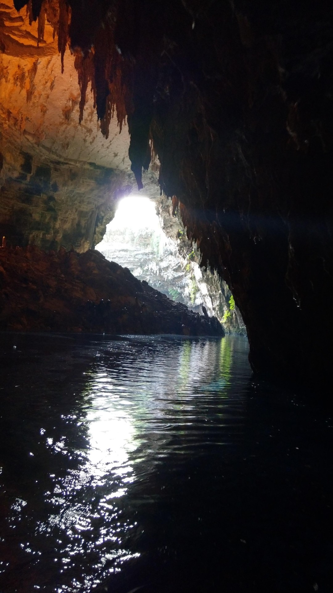 Blick aus einer Höhle entlang eines Baches ins Licht. Von den Felsen hängen Tropfsteine herab. Oben erscheint der Fels teilweise rot m