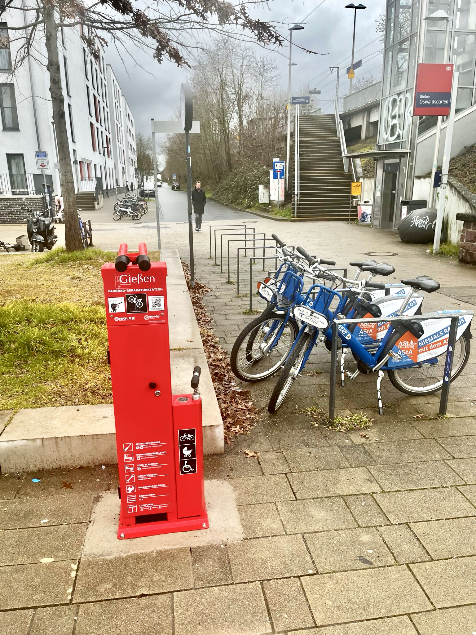 Die rote Reparaturstation in der Größe einer Tanksäule beinhaltet Werkzeuge für eine Fahrrad Reparatur. Daneben parken einige Fahrräder.