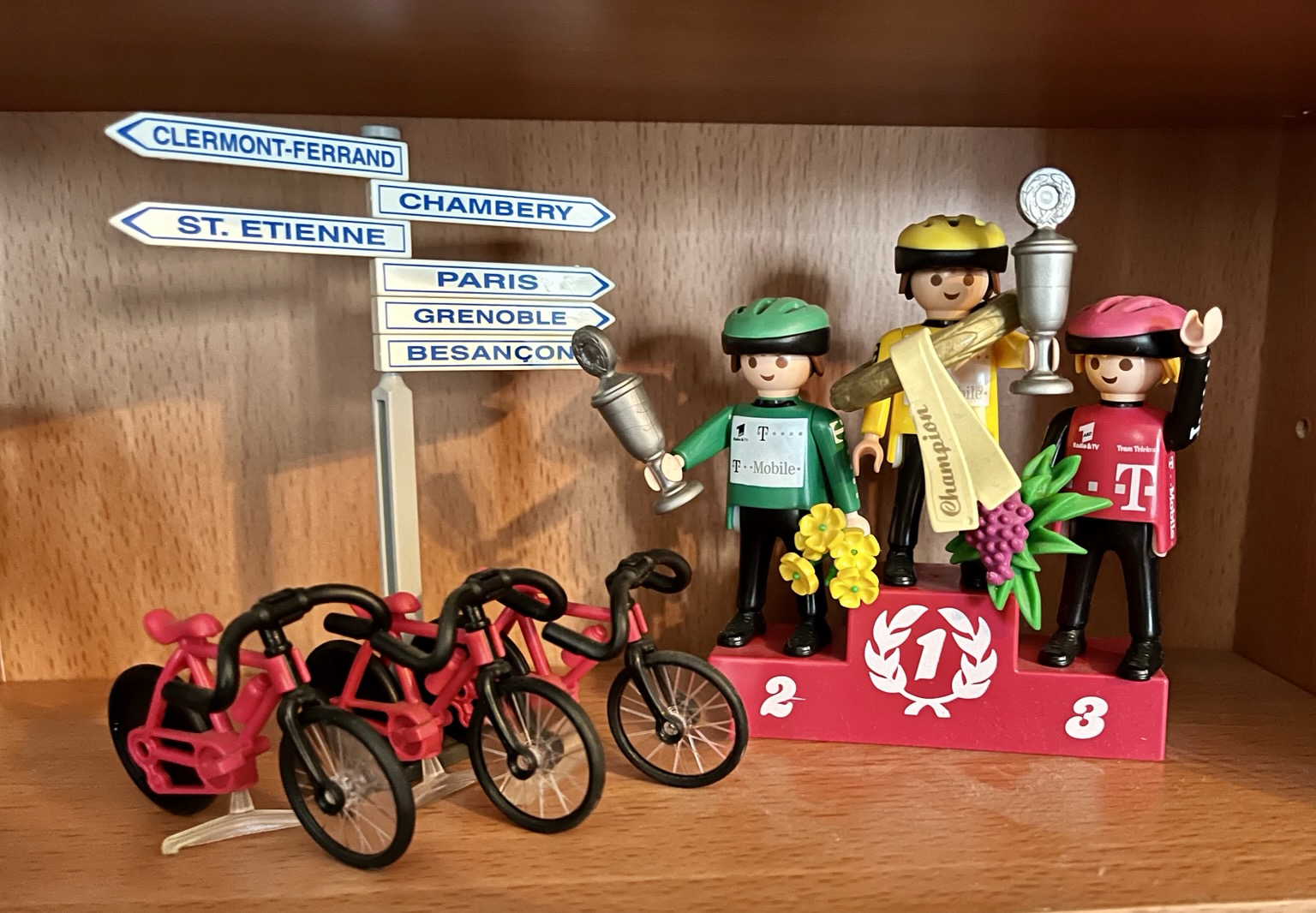 Siegerehrung des Team Telekom bei der Tour de France. Die Sieger mit dem gelben grünen und dem rosa farbenen  Trikot des Team Telekom stehen auf dem Siegerpotest. Vor ihnen ihre drei Fahrräder.. links daneben steht ein Straßenschild mit französischen Ortsnamen.