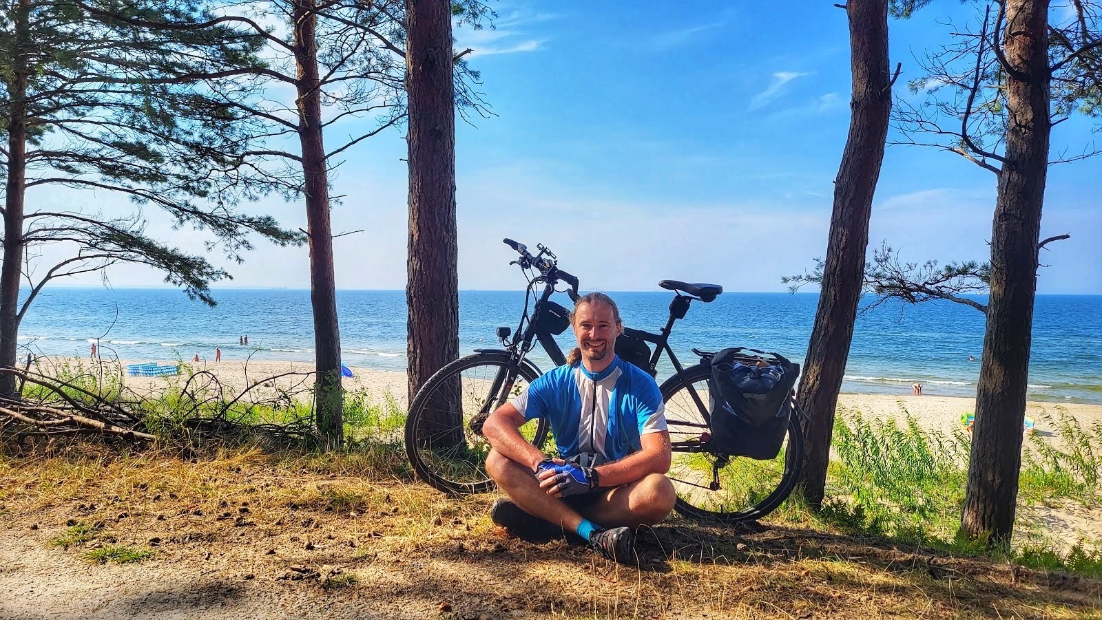 Ich sitze im Halbschatten unter Bäumen vor meinem Fahrrad. Hinter mir ist der Strand und die Ostsee zu sehen.
Es ist sonnig