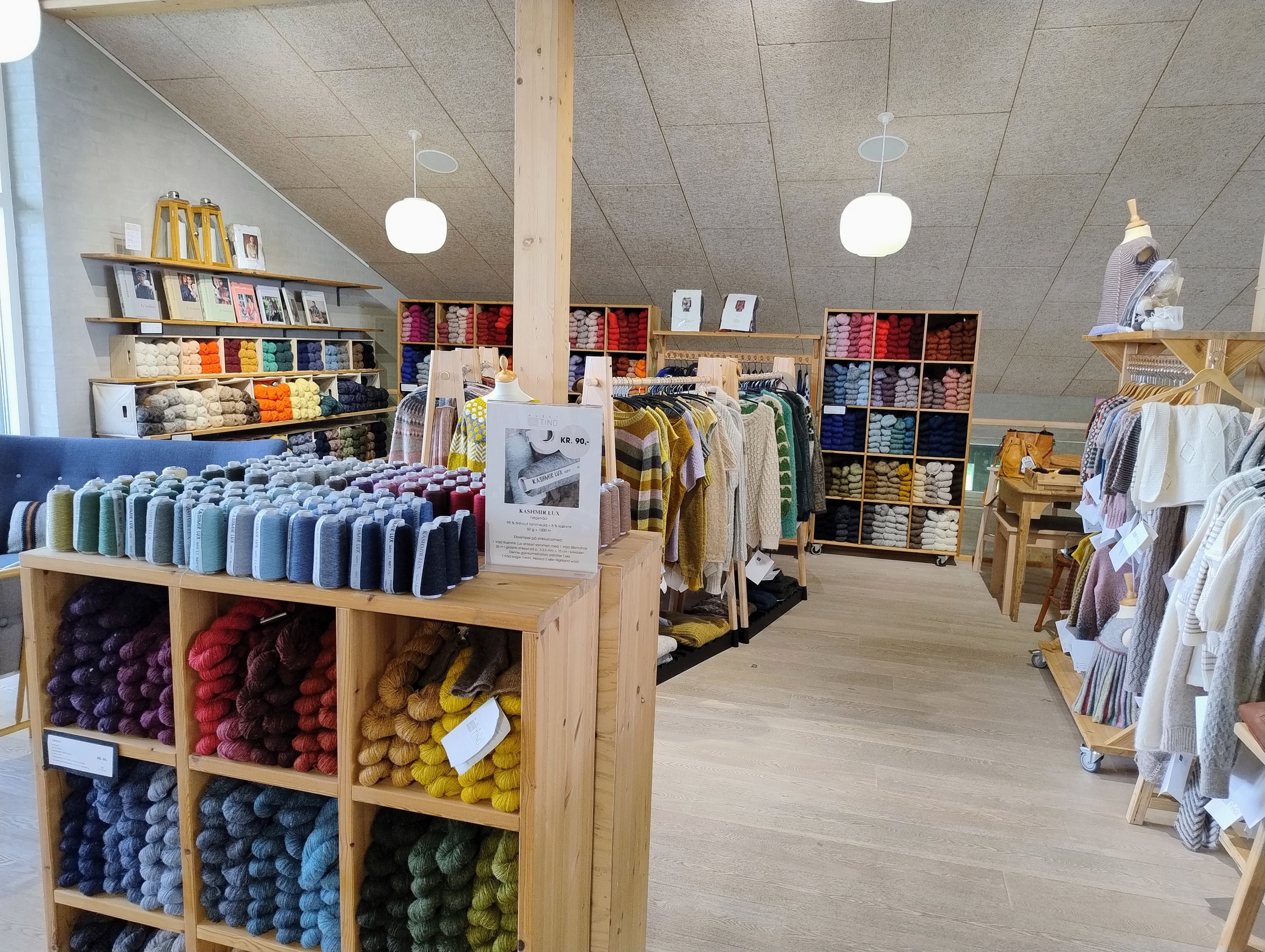 Ein gemütliches Handarbeitsgeschäft - helles Holz, viel Platz, Regale voll Knäuel sortiert im Farbverlauf, mehrere Kleiderständer mit Modellen.