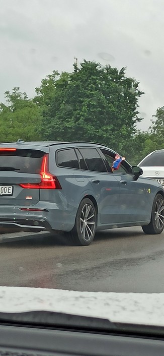 Beifahrer hält eine blaurote Kasperlefigur aus dem Beifahrerfenster eines grauen Volvo Kombi