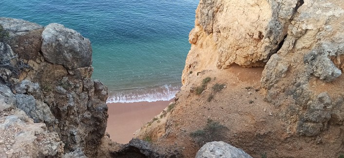 Von oben zwischen Felsen auf einen Sandstrand fotografiert. Wasser türkis und glasklar, leichter Wellengang, Wasser in 15 Meter vom Ufer ins dunkelblau übergeben. Felsen rechts und links grau bis ockerfarben.