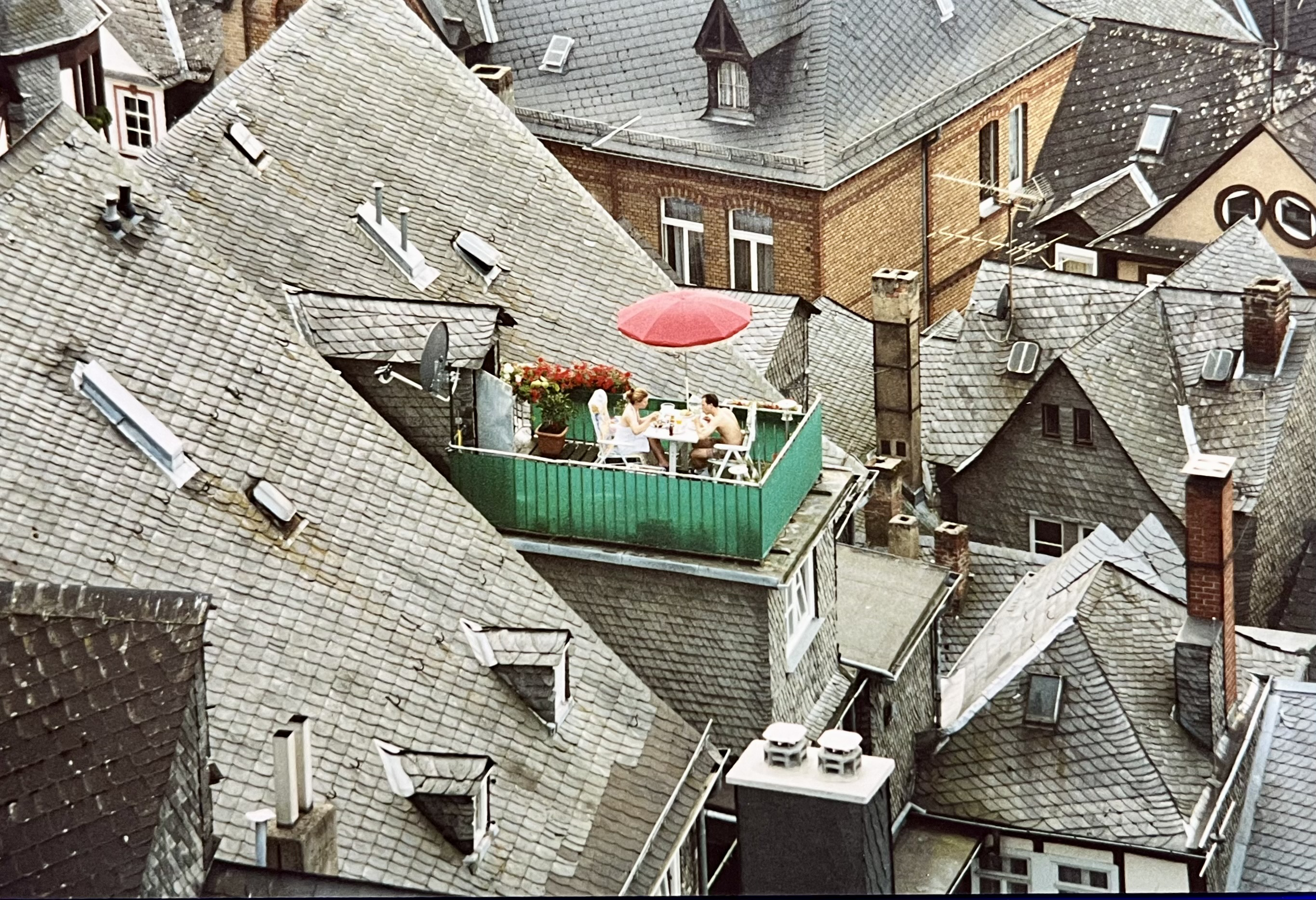Zwischen vielen grauen Dächern sieht man eine Dachterrasse, die von einen grünen Zaun umgeben ist. Darauf sitzen zwei Menschen an einen Tisch unter einen roten Schirm und essen. Zwischen vielen grauen Dächern sieht man eine Dachterrasse, die von einen grünen Zaun umgeben ist. Darauf sitzen zwei Menschen an einen Tisch unter einen roten Schirm und essen.