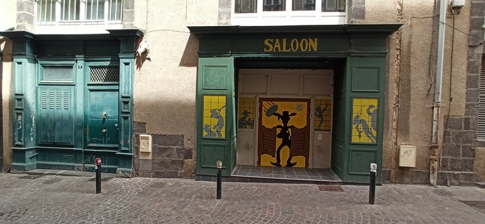 Zwei Holzverkleidungen um die Türen zweier Läden. Die linke ist hellblau/türkis gestrichen, die Rechte grün und die Schrift "Saloon" über der Tür. Auf den zwei Tür Hälften ist die Comicfigur Lucky Luke gemalt, wie er gerade die Hälften einer Saloon Tür teilt und von der Dunkelheit ins Licht tritt