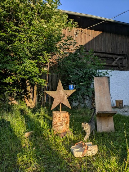 ein großer Holzstern, ein Steckstuhl und Holzschuhe vor einem blühenden Holunder und einer alten Scheune 