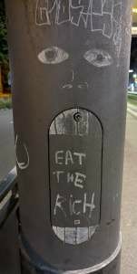Laternenmast mit Bemalung. Augen über einer Klappe als aufgerissser Mund mit Text: eat the rich