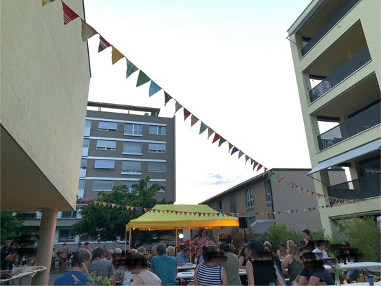 Menschen an Festbänken am Diskutieren. Links und rechts ein Haus, im Hintergrund ein älteres Hochhaus. Ein gelbes Sonnensegel.