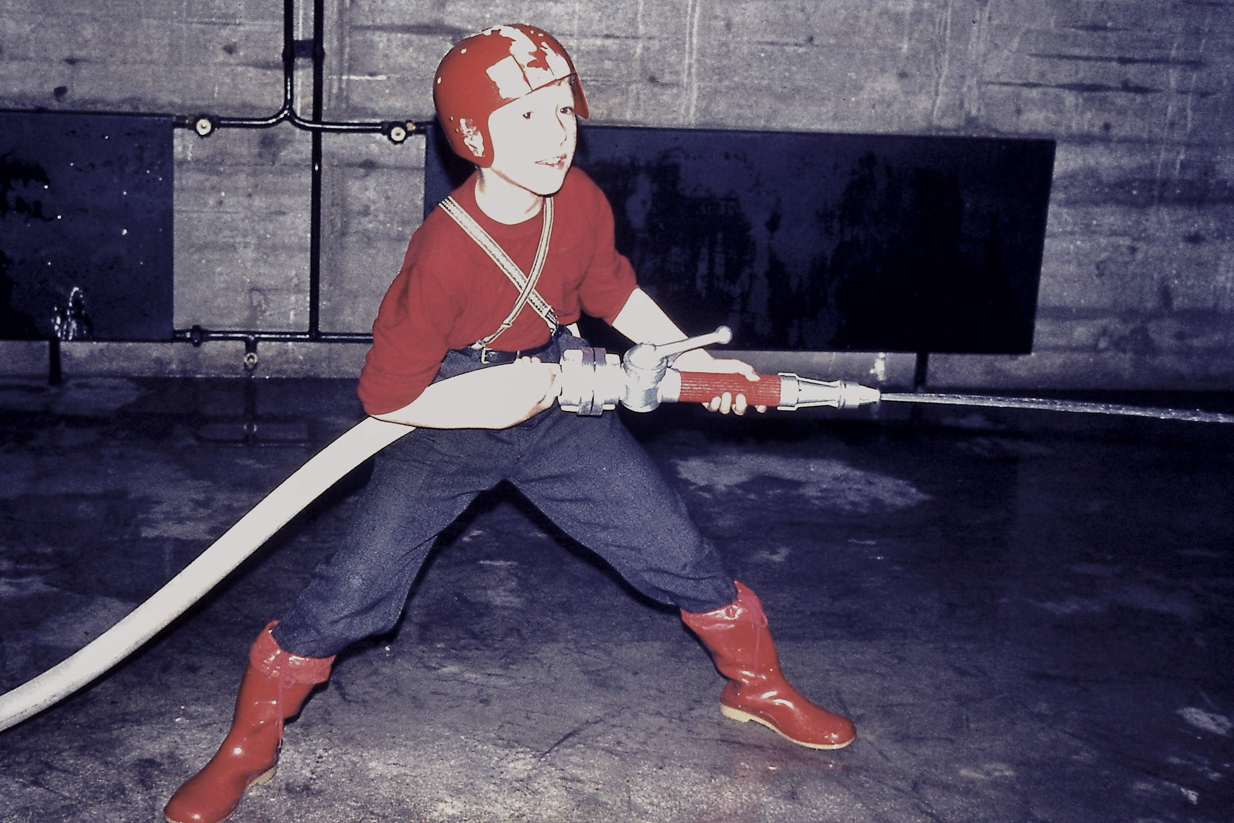 Ein Junge mit roten Helm,roten Pulli und roten Stiefeln hält breitbeinig einen Spritzenden Feuerwehrschlauch in den Händen. Ein Junge mit roten Helm,roten Pulli und roten Stiefeln hält breitbeinig einen Spritzenden Feuerwehrschlauch in den Händen.
