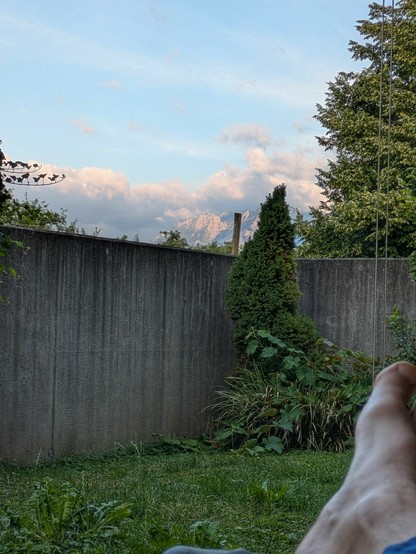 rechts im Vordergrund ein nackter Fußnhinter der Betonwand Berge und Wolken und blauer Himmel 