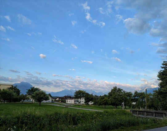 Wiese, Häuser und entfernte Berge und blauer Himmel drüber 