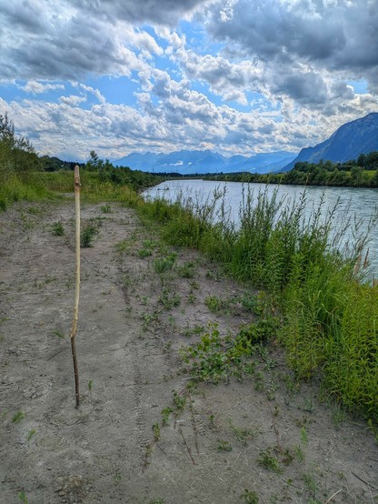 ein grünliches Gewässer, blauer Himmel mit weißen Wolken, ein Stecken steht im Sand vom Ufer,nim Hintergrund schemenhaft Berge