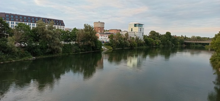 Foto von einer  Brücke über der Donau aufgenommen zeigt es den nördlichen Teil in Richtung Osten. Donau ca. 75 Meter breit, am Ufer links ein weißes langes Gebäude mit schwarzem Dach, dann in ziegelrot ein Alter runder Festungsturm, danach ein neuer Turm in weiß und ultra modern.
