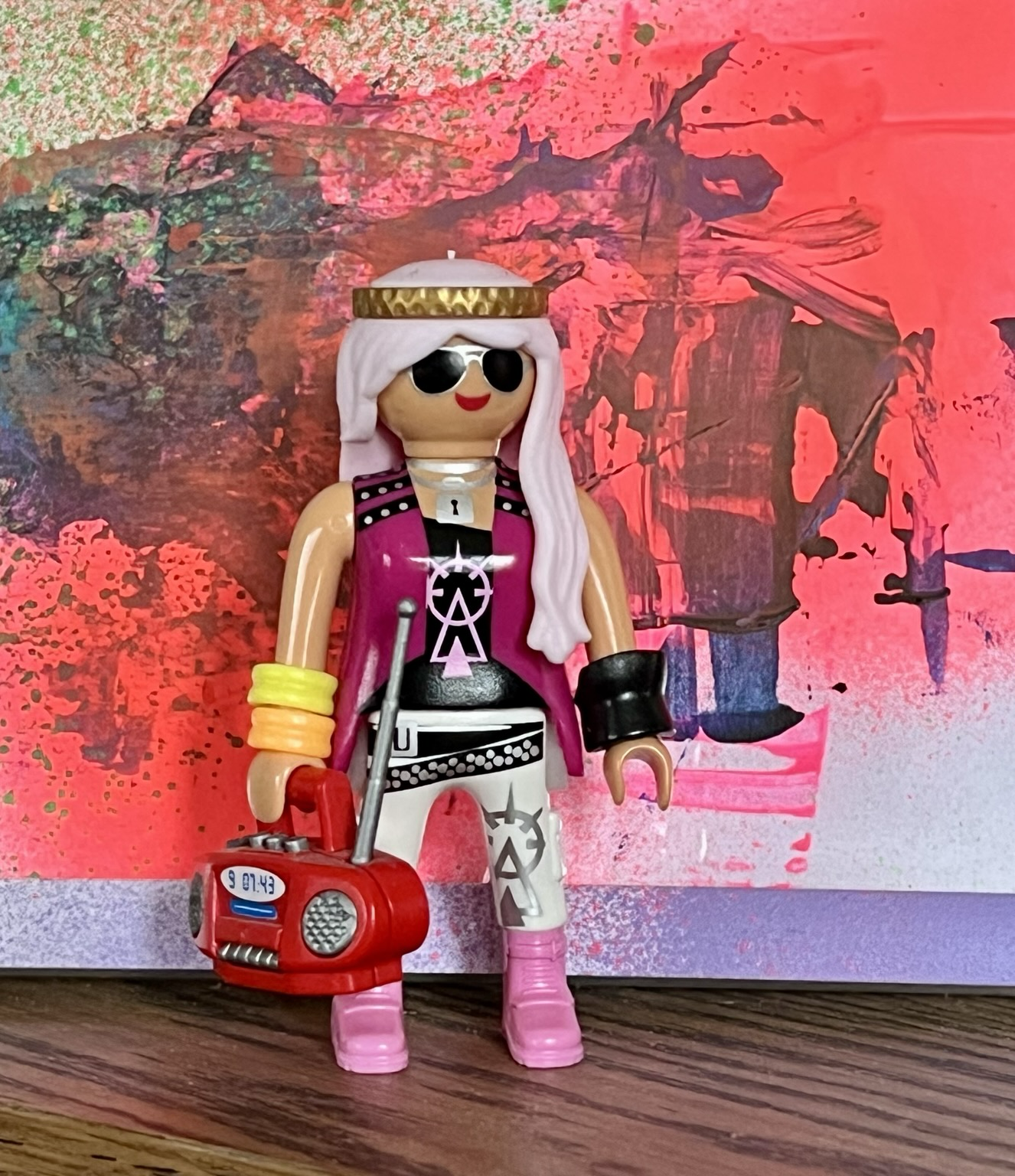 Playmobilfigur. Ein Hippie Girl mit langen hell rosa Haaren, rosaroten Shirt und Rosa schuhen. In der rechten Hand hält sie einen roten Ghettoblaster. Sie steht vor einer Wand mit einem bunten Graffiti Playmobilfigur. Ein Hippie Girl mit langen hell rosa Haaren, rosaroten Shirt und Rosa schuhen. In der rechten Hand hält sie einen roten Ghettoblaster. Sie steht vor einer Wand mit einem bunten Graffiti