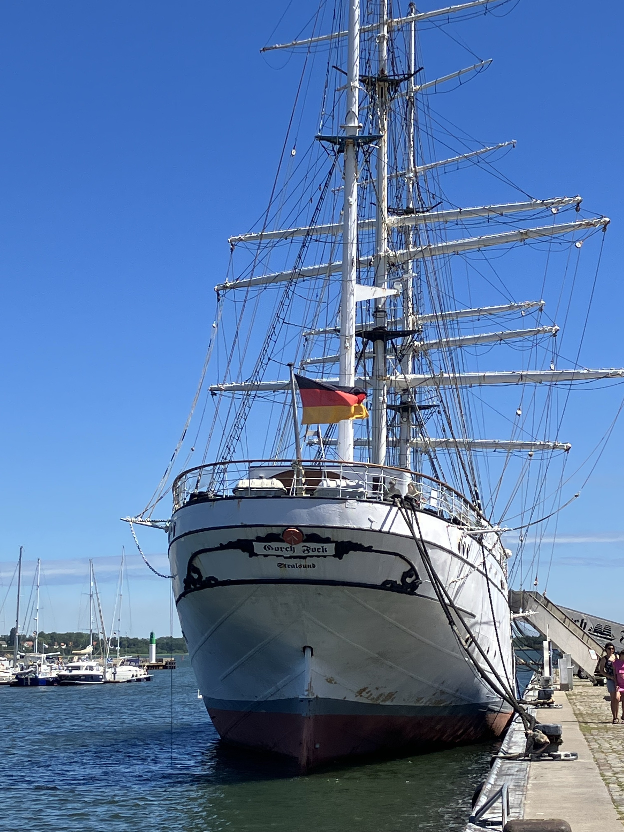 Ein großes Segelschiff, das am Bug die Aufschrift Gorch Fock trägt und in einem Hafen mit einem Hafen angedockt ist. Das Schiff verfügt über mehrere Segel und eine deutsche Flagge. Eine kleine Gruppe von Menschen befindet sich auf dem nahe gelegenen Gehweg. Segelboote sind im Hintergrund zu sehen. Der Himmel ist strahlend blau und wolkenlos. Ein großes Segelschiff, das am Bug die Aufschrift Gorch Fock trägt und in einem Hafen mit einem Hafen angedockt ist. Das Schiff verfügt über mehrere Segel und eine deutsche Flagge. Eine kleine Gruppe von Menschen befindet sich auf dem nahe gelegenen Gehweg. Segelboote sind im Hintergrund zu sehen. Der Himmel ist strahlend blau und wolkenlos.