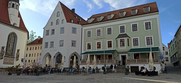 Feier Platz mit mit Bestuhlung von Gasthäusern, Sonnenschirme zusammengeklappt. Das eine Haus hat unten eine Galerie, ebenfalls mit Bestuhlung, ist zweistöckig und lindgrün gestrichen Eines der sechs Fenster im ersten Stock ist als Erker ausgeführt. Das Haus beherberge früher mal den Gasthof Laubenbräu, der als Schriftzug das Gebäude bezeichnet. Links davon ein weißes höheres Haus mit Giebelfront und Laubengängen. Ganz links ein Kirchengebäude mit einem Fenster, das von zwei Heiligenfiguren fla…