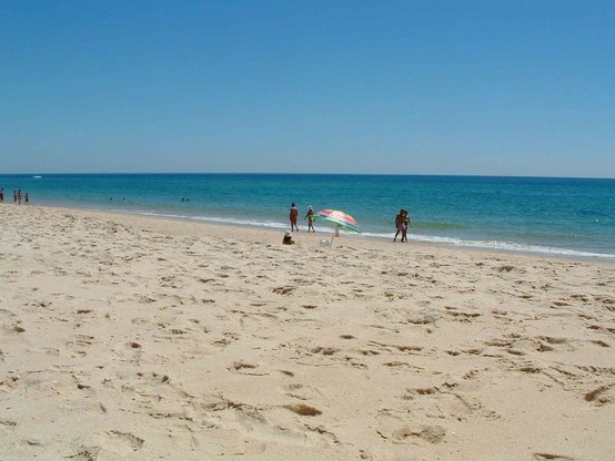 Sandstrand mit vielen Fußabdrücken im Vordergrund, die Uferlinie zieht sich von rechts unten bis links oben durchs Bild. In der Mitte des Bildes und fast am Ufer ein einsamer bunter Sonnenschirm und vier Personen, die am Ufer flanieren. Der Himmel ist blau, es ist MIttagszeit und das Wasser dunkeltürkis. 