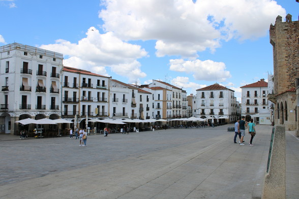 Hauptplatz der Stadt Caceres in Spanien. Rechts ein Turm in ockerfarbenem Granit mit Zinnen oben, dann der großflächige Platz 200x100 Meter, den viele weißgestrichene Häuser umrahmen. Alle bodentiefe Fenster sind mit kleinem Balkon versehen, einige verschatten das Fenster von außen mit einer Markise. Unten auf dem Platz stehen große weiße Sonnenschirme und Bestuhlung vor den Restaurants. Bild Nachmittags um ca. 14 Uhr aufgenommen, blauer Himmel mit weißen Haufenwolken.