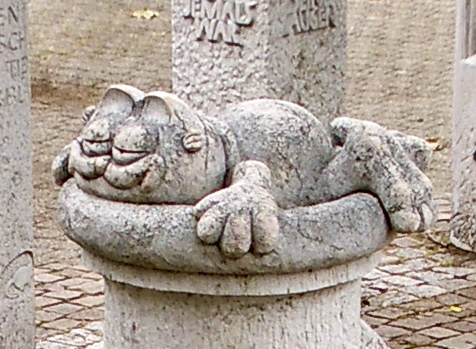 In Weißkrautstein gehauen, liegt der besagte Kater faul, alle viere von sich streckend, auf den runden Oberteil einer Steinsäule In Weißkrautstein gehauen, liegt der besagte Kater faul, alle viere von sich streckend, auf den runden Oberteil einer Steinsäule
