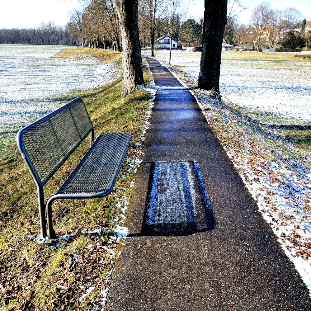 der blick geht einen schmalen asphaltierten fußweg entlang, der oben auf einem kleinen wall oder deich verläuft und wie eine kleine allee beidseitig von bäumen gesäumt ist. links und rechts des walls liegen agrarische wiesen auf deren kurzem bewuchs eine dünne schicht schnee liegt. ganz hinten im bild beginnt der auwald. im vordergrund steht links neben dem weg eine parkbank aus stahlrohr und drahtgitter. die wirft einen schatten auf den weg und genau da wo der schatten ist bzw. vor einer weile war liegt noch etwas raureif oder schnee, während er sonst überall dort wo die sonne hinkommt getaut ist. der blick geht einen schmalen asphaltierten fußweg entlang, der oben auf einem kleinen wall oder deich verläuft und wie eine kleine allee beidseitig von bäumen gesäumt ist. links und rechts des walls liegen agrarische wiesen auf deren kurzem bewuchs eine dünne schicht schnee liegt. ganz hinten im bild beginnt der auwald. im vordergrund steht links neben dem weg eine parkbank aus stahlrohr und drahtgitter. die wirft einen schatten auf den weg und genau da wo der schatten ist bzw. vor einer weile war liegt noch etwas raureif oder schnee, während er sonst überall dort wo die sonne hinkommt getaut ist.