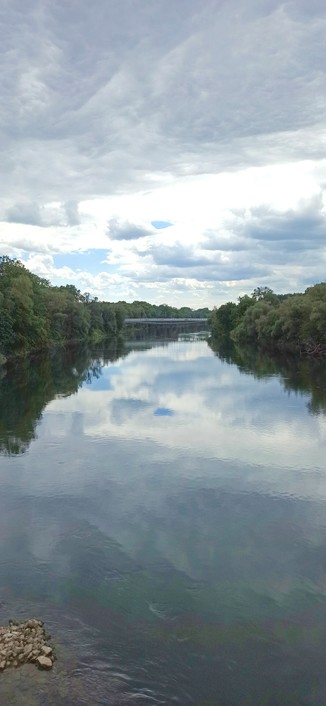Fluß von der der Mitte der Donaubrücke nach Westen fotografiert. Himmel stark bewölkt mit einzelnen blauen Flecken, spiegelt sich alles in der ruhig dahinfließenden Donau. In der Ferne ist die andere Brücke über die Donaus zu sehen und die hellen senkracht verlaufenden Stäbe des Brückengeländers.