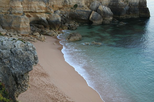 kleine Wellen plätschern an einen dunkelockerfarbenen Sandstrand, der von Felsen gerahmt ist. Wasser ist leicht türkis, die Sonne nicht aufgegangen. Es finden sich Fußspuren der Busucher am Vortag im Sand. 