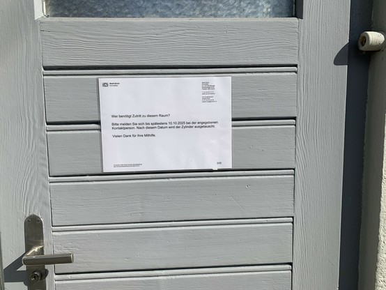Aufnahme einer teilweise erfassten, grau gestrichenen Tür von aussen, im oberen Teil ein Milchglasfenster. Die Tür ist rechts angeschlagen und geht nach aussen auf. Links ist gerade noch die Türfalle mit Schloss sichtbar. Unterhalb des Fensters ein laminiertes Plakat in A4 quer mit dem Briefkopf der Immobilienverwaltung der Stadt Zürich. Man bittet interessierte Personen, sich zu melden, ansonsten der Schliesszylinder ab 10.10. ausgetauscht werde.