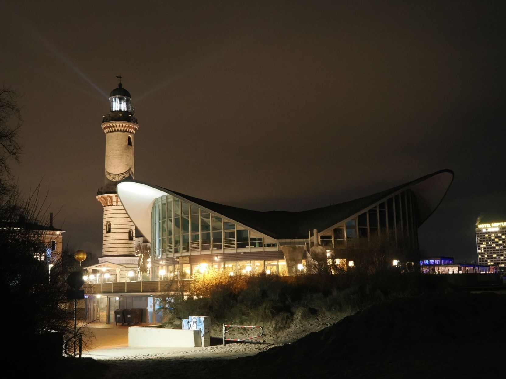 Eine Nachtaufnahme. Im linken Drittel ist ein Leuchtturm zu sehen, davor steht in den rechten zwei Dritteln ein großes Gebäude mit Glaswänden und einem geschwungenen Dach. Vom Leuchtturm aus sind nach rechts und links jeweils ein Lichtstrahl zu sehen. Eine Nachtaufnahme. Im linken Drittel ist ein Leuchtturm zu sehen, davor steht in den rechten zwei Dritteln ein großes Gebäude mit Glaswänden und einem geschwungenen Dach. Vom Leuchtturm aus sind nach rechts und links jeweils ein Lichtstrahl zu sehen.