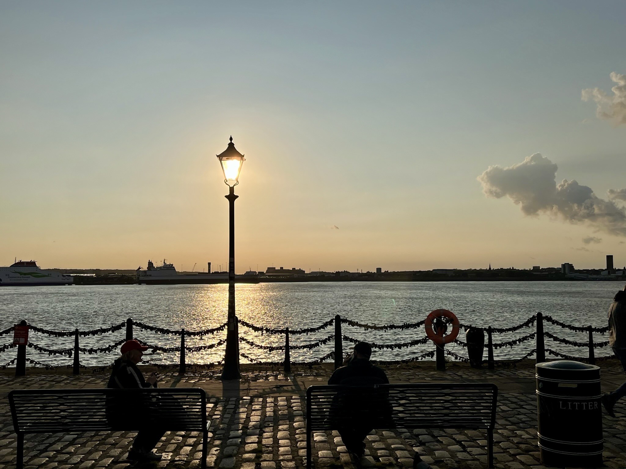 Eine Szene am Wasser während des Sonnenuntergangs, mit einem Laternenpfahl.Die Sonne ist direkt hinter der Laterne. Es sieht aus, als sei sie elektrisch beleuchtet.
Silhouetten von Menschen, die auf Bänken sitzen, und entfernten Booten auf dem Wasser. Ein Rettungsring ist neben einer Barriere sichtbar, mit einer Skyline der Stadt im Hintergrund. Eine Szene am Wasser während des Sonnenuntergangs, mit einem Laternenpfahl.Die Sonne ist direkt hinter der Laterne. Es sieht aus, als sei sie elektrisch beleuchtet.
Silhouetten von Menschen, die auf Bänken sitzen, und entfernten Booten auf dem Wasser. Ein Rettungsring ist neben einer Barriere sichtbar, mit einer Skyline der Stadt im Hintergrund.