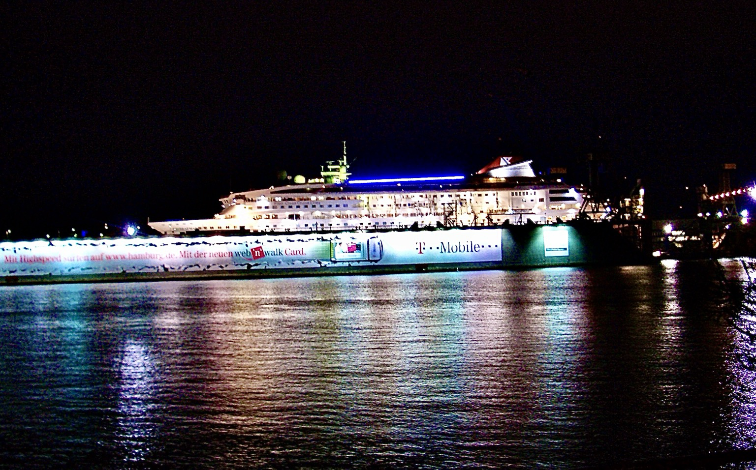 Es ist Nacht. In einen hell und bunt beleuchteten Dock steht ein Passagierschiff. Im Vordergrund sieht man schwarzes Wasser, auf dem sich viele bunte Lichter spiegeln Es ist Nacht. In einen hell und bunt beleuchteten Dock steht ein Passagierschiff. Im Vordergrund sieht man schwarzes Wasser, auf dem sich viele bunte Lichter spiegeln