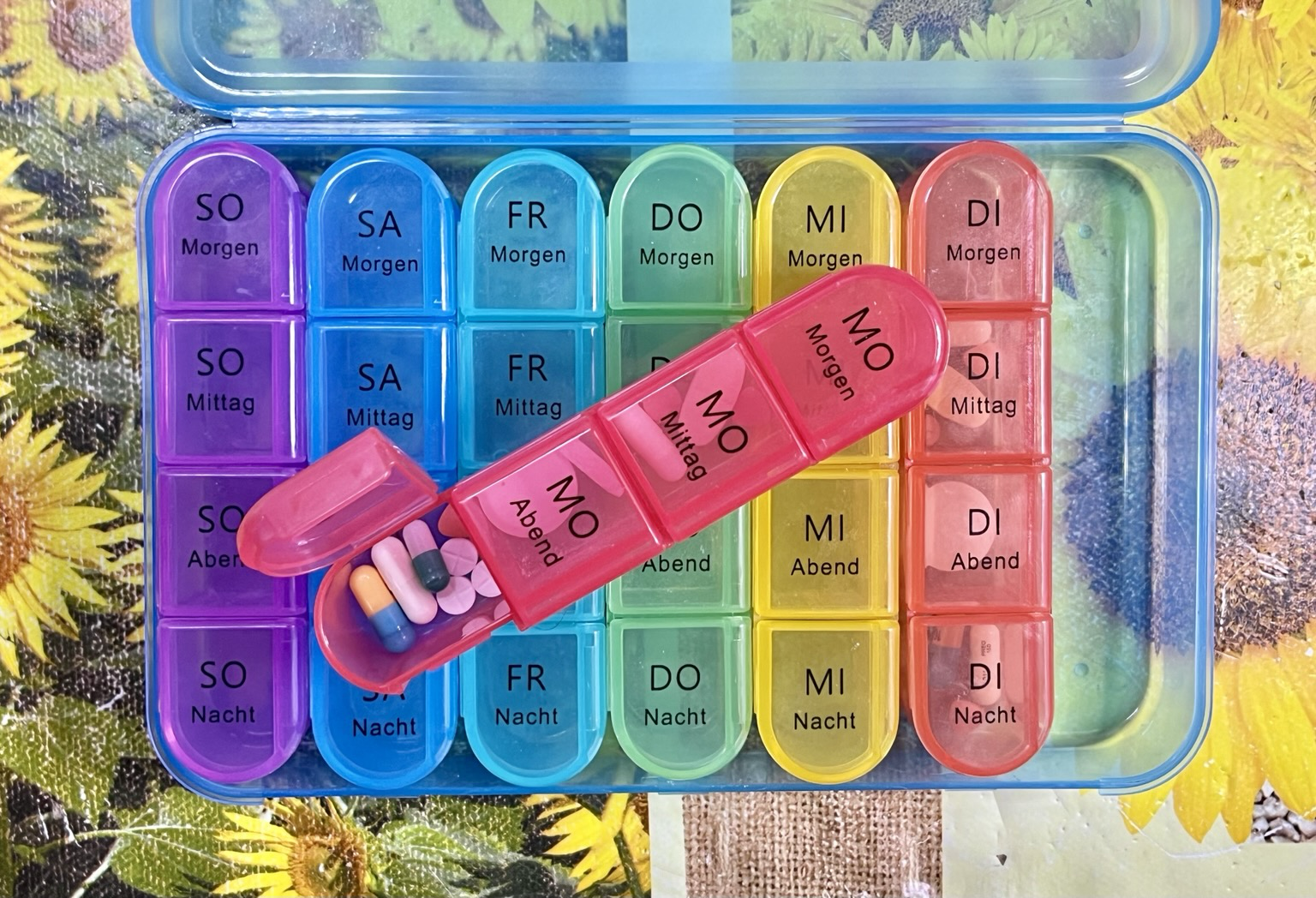 Das Bild zeigt eine farbenfrohe Medikamentenbox mit Tages- und Nachtsegmenten, die in verschiedenen Farben kodiert sind. Die Box ist auf einem Tisch mit einem Sonnenblumenmuster platziert. Die Tagessegmente sind mit den deutschen Wörtern "Morgen," "Mittag" und "Abend" beschriftet, während die Nachtsegmente "Nacht" bezeichnen. Die Box ist in sieben Farben unterteilt, jeweils für einen Wochentag: Sonntag (SO), Samstag (SA), Freitag (FR), Donnerstag (DO), Mittwoch (MI) und Dienstag (DI). Ein roter Behälter mit den Wörtern "MO" (Montag) und "DI" (Dienstag) liegt auf der Box und enthält verschiedene Pillen. Die Farben der Segmenten sind eindeutig und erleichtern die Identifikation der richtigen Zeit für die Medikamente Das Bild zeigt eine farbenfrohe Medikamentenbox mit Tages- und Nachtsegmenten, die in verschiedenen Farben kodiert sind. Die Box ist auf einem Tisch mit einem Sonnenblumenmuster platziert. Die Tagessegmente sind mit den deutschen Wörtern "Morgen," "Mittag" und "Abend" beschriftet, während die Nachtsegmente "Nacht" bezeichnen. Die Box ist in sieben Farben unterteilt, jeweils für einen Wochentag: Sonntag (SO), Samstag (SA), Freitag (FR), Donnerstag (DO), Mittwoch (MI) und Dienstag (DI). Ein roter Behälter mit den Wörtern "MO" (Montag) und "DI" (Dienstag) liegt auf der Box und enthält verschiedene Pillen. Die Farben der Segmenten sind eindeutig und erleichtern die Identifikation der richtigen Zeit für die Medikamente