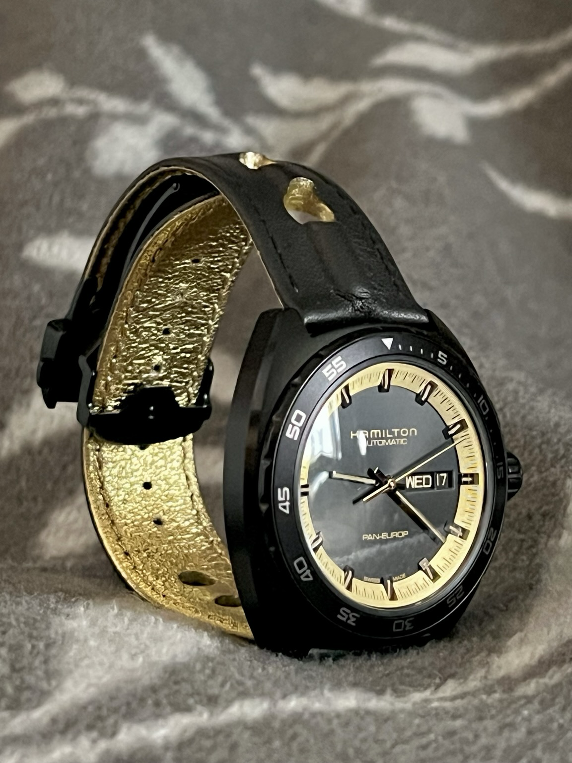 Eine Armbanduhr mit schwarzem Gehäuse und Armband, dass auf der Innenseite mit glänzenden Gold belegt ist. Das schwarze Zifferblatt ist von einem goldenen Kreis mit den Stunden-und Minutenstrichen umgeben. Auf den anderen Bildern ist die Uhr aus anderen Perspektiven zu sehen, unter anderem von hinten, wo man durch Glas hindurch das Automatik-Uhrwerk sehen kann