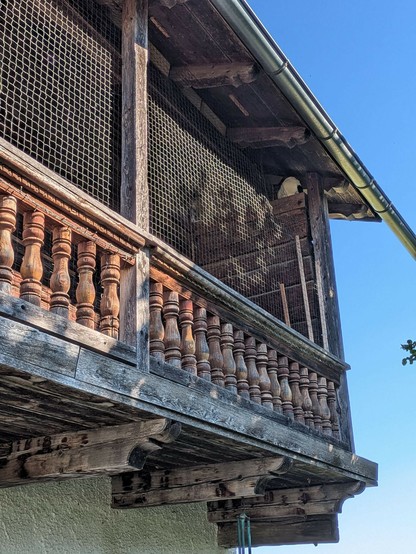 ganz oben rechts in einem Holzbalkon unter dem Dach sitzt eine kleine fast weiße Katze