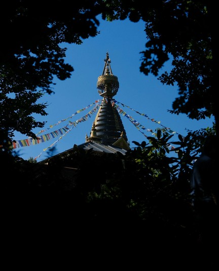 der Turm eines Stupa mit bunten Gebetsfahnen vor blauem Himmel und eingerahmt von Laubbäumen