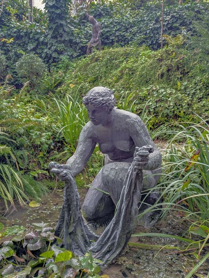 ein Mädchen in Bronze, das am Wasser Wäsche wäscht und im Hintergrund ein Schmied in Bronze