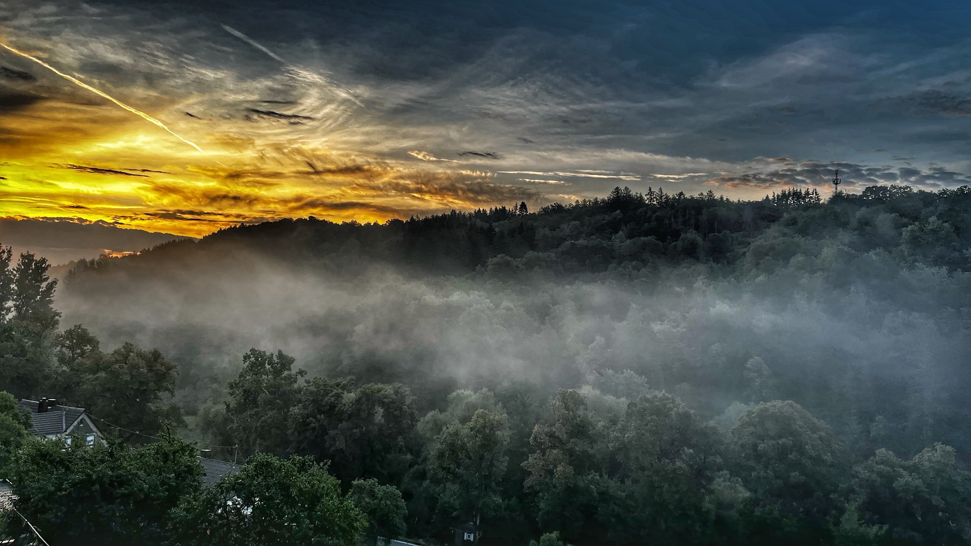 Ein malerischer Blick auf einen Nebelwald bei Sonnenaufgang, mit einem leuchtend orangefarbenen und gelben Himmel mit verstreuten Wolken. Dichte Bäume sind im Vordergrund sichtbar, teilweise durch Nebel verdeckt. Ein malerischer Blick auf einen Nebelwald bei Sonnenaufgang, mit einem leuchtend orangefarbenen und gelben Himmel mit verstreuten Wolken. Dichte Bäume sind im Vordergrund sichtbar, teilweise durch Nebel verdeckt.
