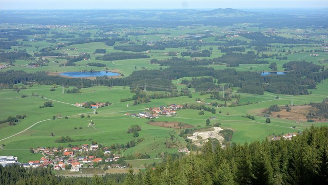 Blick in Tal auf eine Ortschaft, auf Wiesen, die von kleinen sattgrünen Nadelwäldchen unterbrochen werden. Hinter der ersten quer durchs Bild laufende Linie von Wäldchen leuchten zwei kleine strahlend blaue Seen. Im Hintergrund in Richtung Himmel wird es dunstig und verschwimmt mit dem blauen Himmel. 
