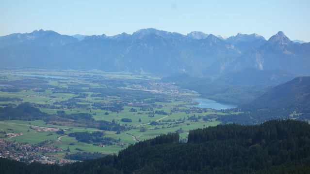 Bergpanorama mit zwei Seen und ganz klein das Schloß Neuschwanstein, davor im Panorama viele grüne wiesen, unterbrochen von Wäldchen und kleinen Weilern.