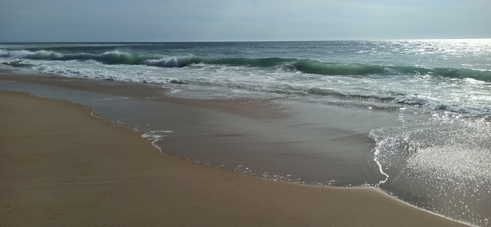 Wellen laufen auf einen flachen Sandstrand, die späte Nachmittagssonne bringt das Wasser zum Glitzern. Das Bild zeigt die Zone, auf der das Wasser schäumend auf den Sand läuft und die Wellen beim kleiner werden sich kreuzen.Himmel ist blau, der Sand dunkles ocker. 