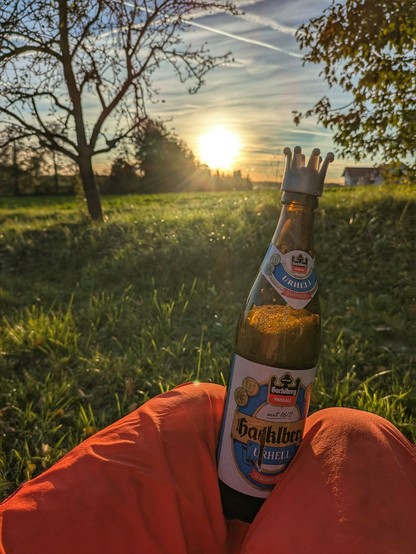 eine Bierflasche mit einem Verschluss in Form einer Krone klemmt zwischen Beinen in einer orangen Hose.nDahinter eine niedrige Sonne am milchigblauen Himmel