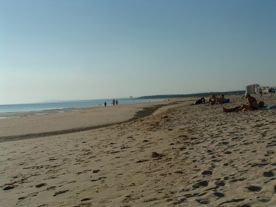 Sandstrand auf linker Seite des Bilds unter blauem Himmel, rechts das Meer ohne Wellen. Einige Leute auf dem Strand, aber nicht überlaufen. Kamera ziemlich ins Gegenlicht der tiefstehenden Spätnachmittagssonne gehalten und abgedrückt. 