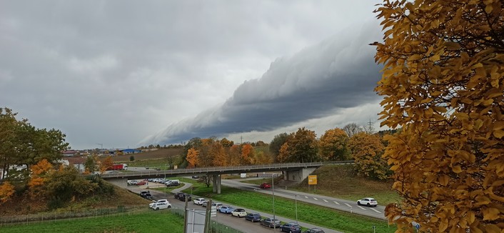 Blick vom 3.Stock auf zwei sich kreuzende Bundesstraßen, die eine wir mit Brücke über die andere geführt. Bäume im Bild herbstlich gelb und Rot gefärbt. Am Himmel eine dunkelgraue Wolkenwalze durch das ganze Bild parallel zu einer der Straßen.