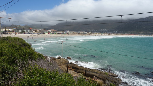 Meeresbucht, an den Strand schließt eine Stadt an. Das Meer läuft auf sehr flachen Sandstrand, es sind viele Bedende am Meer. Das Wasser ist tiefgrün-türkis und läuftmit langen schäumenden Wellenkämmen den Strand hinauf. Bild leicht dunstig, über den Hügeln im Hintergrund liegt das sogenannte Blanket, das man bei besonderen Wetterlangen auf fast allen Hügeln der Cape Region sehen können und bedeutet, eine dichte Wolkendecke liegt satt und weiß auf dem Hügel.